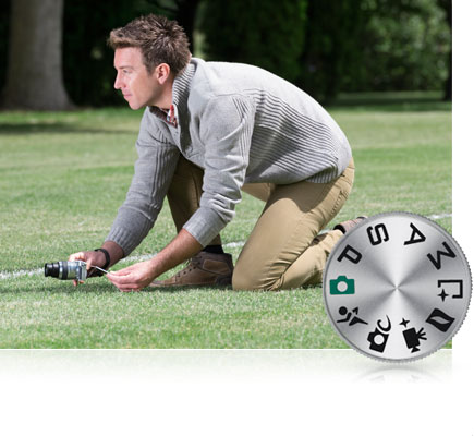 Photo of a man kneeling on the grass shooting at a low angle using the tilting touchscreen of the Nikon 1 J5 inset with the mode dial