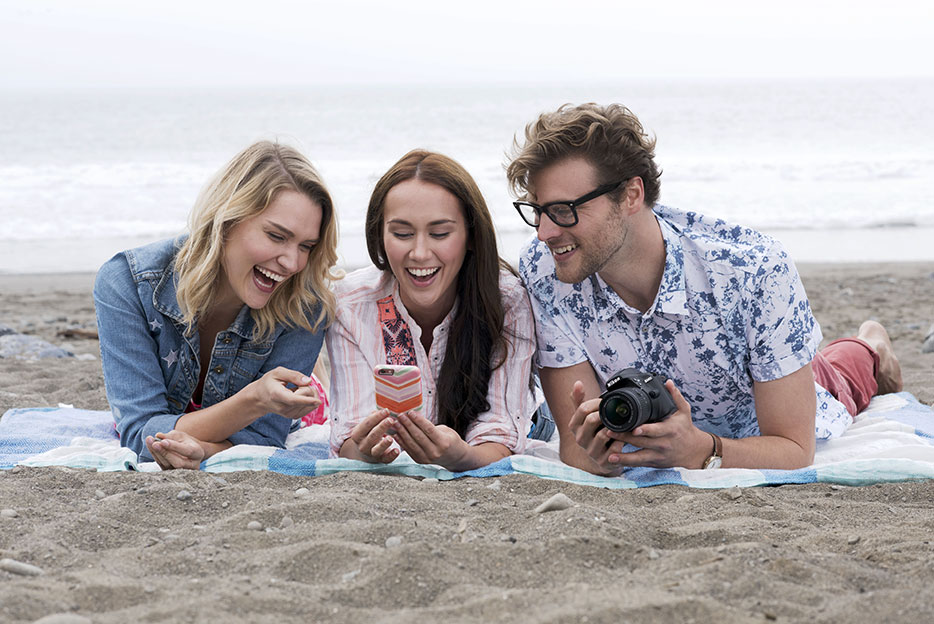 Nikon D3400 photo of two women and a man on a beach blanket looking at photos on a smartphone