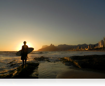 Photo of the sun setting over Rio De Janeiro, Brazil while a surfer stands in the shore break