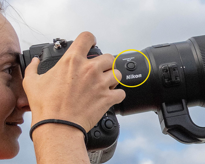 Photo of a woman looking through the viewfinder of a camera with the NIKKOR Z 800mm f/6.3 VR S lens attached, with the memory set button circled in yellow