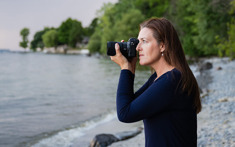 photo of a woman shooting with the NIKKOR Z 17-28mm f/2.8 lens on a camera, outdoors by a lake