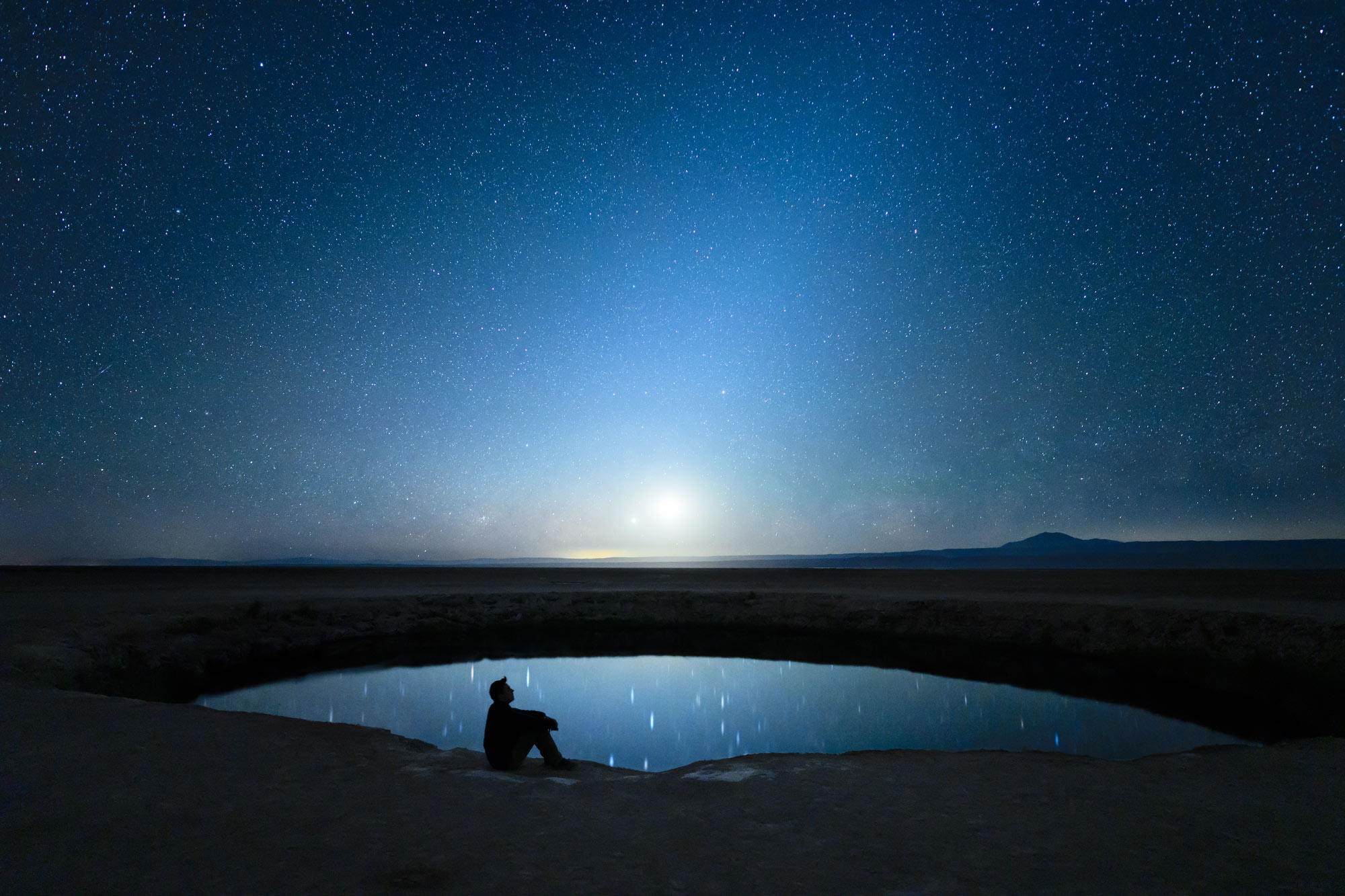 Foto notturna di un cielo stellato e del riflesso in uno stagno, con una figura in silhouette che fissa il cielo, scattata con il NIKKOR Z 20mm f/1.8 S